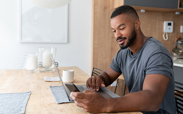 man-sitting-at-table-using-laptop-and-looking-at-phone-join-hollister-consumer-services